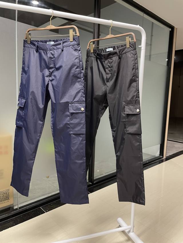 普拉达2023Spra*A这款工装长裤采用re-Nylon再生尼龙面料打造，饰以标志性的涂珐琅三角形金属徽标。再生尼龙纱线econyl通过提纯海洋塑料、彰显品牌
